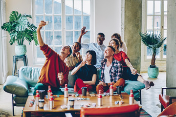 Satisfied hipster guys smiling at smartphone front camera while clicking selfie content via modern application for sharing media content, happy diverse friends enjoying leisure weekend for card gaming