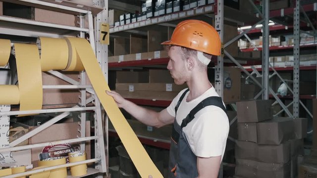 A factory worker wearing a helmet is measuring the tape. Then he is rolling it back.