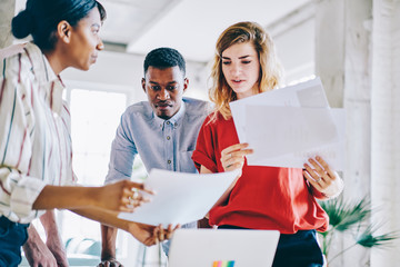 Obraz premium Group of multiracial male and female colleagues talking near table with laptop technology and papers preparing to university exams together, hipster guys discussing ideas during gather cooperation