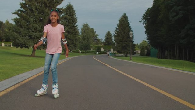 Carefree positive adorable black teenage girl with trendy haircut roller skating in public park. Joyful mixed race preadolescent child having fun, enjoying rollerblading outdoors on summer day.