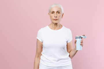 Beautiful elderly gray-haired female woman 60s 70s wearing white blank empty casual t-shirt posing hold bottle of water looking camera isolated on pastel pink color wall background studio portrait.