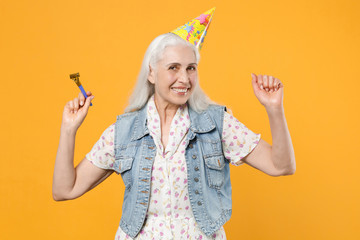 Obraz premium Smiling elderly gray-haired female woman 60s 70s wearing casual dress denim waistcoat birthday hat celebrating hold pipe dancing clenching fists isolated on yellow color background studio portrait.