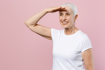 Smiling elderly gray-haired female woman 60s 70s wearing white blank casual t-shirt posing holding hand at forehead looking far away distance isolated on pastel pink color background studio portrait.