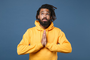 Pleading young african american man in casual yellow streetwear hoodie posing isolated on blue background studio portrait. People sincere emotions lifestyle concept. Holding hands folded in prayer.