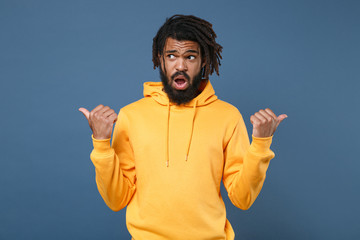 Shocked young african american man guy wearing yellow streetwear hoodie posing isolated on blue wall background studio portrait. People lifestyle concept. Mock up copy space. Pointing thumbs aside.