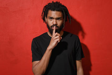 Secret young african american man guy with dreadlocks 20s in black t-shirt posing saying hush be quiet with finger on lips shhh gesture looking camera isolated on red color background studio portrait.