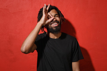 Funny young african american man guy with dreadlocks 20s in black casual t-shirt posing holding hands near eyes, imitating glasses or binoculars isolated on red color wall background studio portrait.