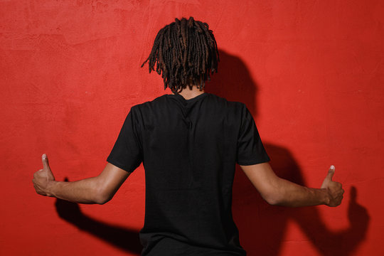 Back Rear View Of Young African American Man Guy With Dreadlocks 20s Wearing Black Casual T-shirt Posing Showing Thumbs Up Spreading Hands Isolated On Bright Red Color Wall Background Studio Portrait.