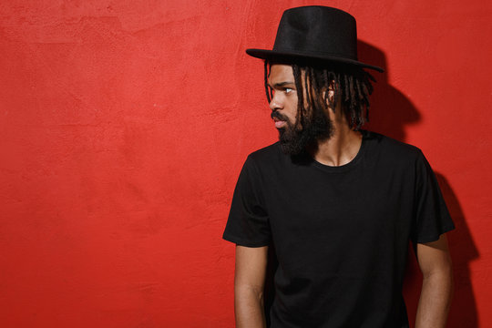 Serious Handsome Attractive Young African American Man Guy With Dreadlocks 20s Wearing Black Casual T-shirt Hat Posing Looking Aside Isolated On Bright Red Color Wall Background Studio Portrait.