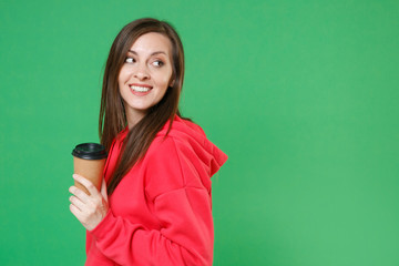Side view of smiling beautiful pretty young brunette woman 20s in red casual streetwear hoodie posing looking aside hold paper cup of coffee or tea isolated on green color background studio portrait.