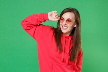 Smiling cheerful attractive beautiful young brunette woman 20s wearing bright red casual streetwear hoodie eyeglasses posing looking aside isolated on green color wall background studio portrait.