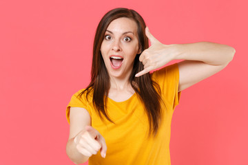 Surprised shocked young brunette woman 20s in yellow casual t-shirt doing phone gesture like says call me back pointing index finger on camera isolated on pink color wall background studio portrait.