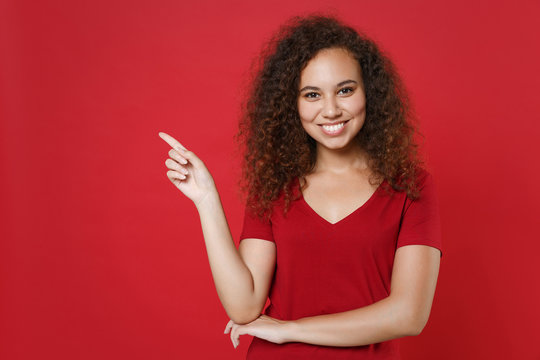 Smiling Young African American Woman Girl In Casual T-shirt Posing Isolated On Red Background Studio Portrait. People Emotions Lifestyle Concept. Mock Up Copy Space. Pointing Index Finger Aside Up.