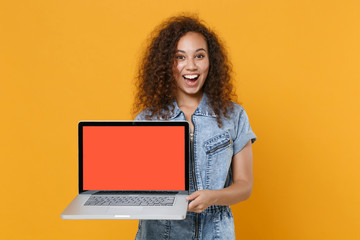 Naklejka premium Surprised young american girl in casual denim clothes isolated on yellow wall background studio portrait. People lifestyle concept. Mock up copy space. Hold laptop pc computer with blank empty screen.