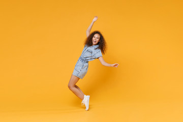 Fototapeta na wymiar Full length portrait of cheerful young african american woman girl in denim clothes isolated on yellow background. People lifestyle concept. Mock up copy space. Dancing standing on toes, rising hands.