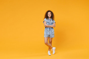Full length portrait of smiling young african american girl in denim clothes isolated on yellow wall background studio portrait. People lifestyle concept. Mock up copy space. Holding hands crossed. - obrazy, fototapety, plakaty