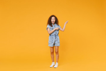 Full length portrait of surprised young african american woman girl in denim clothes isolated on yellow background studio portrait. People emotions lifestyle concept. Pointing index fingers aside.