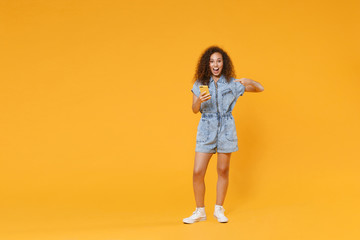 Fototapeta na wymiar Full length portrait of excited young african american woman girl in denim clothes isolated on yellow wall background in studio. People lifestyle concept. Pointing index finger on mobile cell phone.