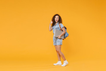 Fototapeta na wymiar Full length portrait funny african american girl student in denim clothes, backpack isolated on yellow background. Education in high school university college. Hold laptop computer, showing thumb up.