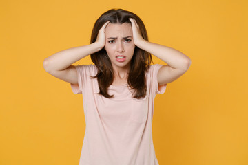 Dissatisfied worried tired young brunette woman wearing pastel pink casual t-shirt standing posing put hands on head having headache isolated on bright yellow color wall background studio portrait.
