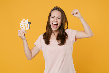 Happy young brunette woman 20s wearing pastel pink casual t-shirt posing hold house bunch of keys doing winner gesture keeping eyes closed isolated on bright yellow color background studio portrait.