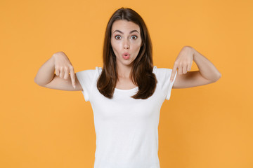 Shocked amazed young brunette woman 20s in white blank empty design casual t-shirt posing pointing index fingers down on mock up copy space isolated on yellow color wall background studio portrait.