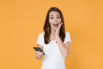 Fototapeta premium Surprised young brunette woman 20s in white blank empty design casual t-shirt using mobile cell typing sms message keeping mouth open put hand on cheek isolated on yellow background studio portrait.