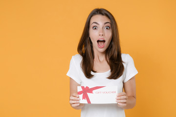 Fototapeta na wymiar Excited surprised shocked young brunette woman 20s wearing white blank casual t-shirt posing hold in hands gift certificate keeping mouth open isolated on yellow color wall background studio portrait.