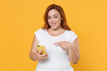 Smiling young redhead plus size body positive female woman girl 20s in white casual t-shirt posing pointing index finger on mobile cell phone isolated on yellow color wall background studio portrait.