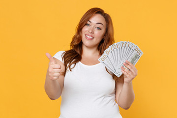 Smiling young redhead plus size body positive female woman girl in white casual t-shirt hold fan of cash money in dollar banknotes showing thumb up isolated on yellow color background studio portrait.