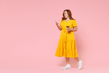 Full length portrait attractive young redhead plus size body positive female woman 20s in yellow dress hold paper cup coffee or tea using mobile phone isolated on pastel pink color background studio.