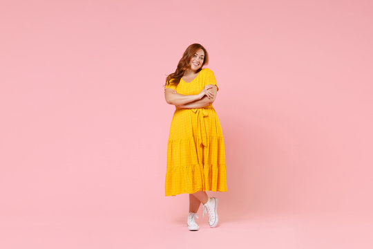 Full length portrait pretty smiling beautiful young redhead plus size body positive female woman girl 20s in yellow dress posing holding hands crossed isolated on pastel pink color background studio.