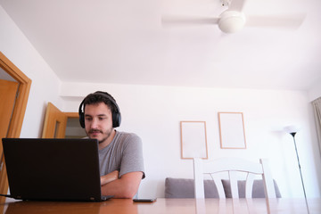 Young man wearing headphones watching series, videos, online classes on a laptop with the fan on. Studying online and e-learning concept.
