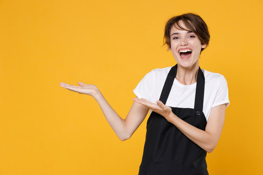 Cheerful Young Female Woman 20s Barista Bartender Barman Employee In White Casual T-shirt Apron Pointing Hands Aside On Mock Up Copy Space Isolated On Yellow Color Wall Background Studio Portrait.