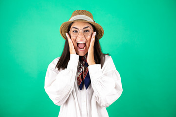 Young beautiful business woman wearing hat over isolated green background with her hands over her mouth and surprised