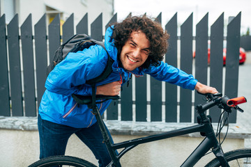 Smiling positive man posing with his bike after bicycling in the street on a rainy day next to the fence's house. Male courier with curly hair in blue raincoat delivers parcel cycling with a bicycle