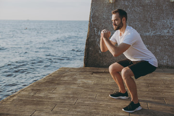 Full length portrait side view of handsome attractive young athletic man guy 20s in white t-shirt black shorts posing training doing exercise squatting looking aside at sunrise over the sea outdoors.