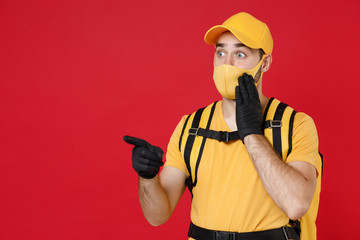 Delivery employee man in yellow cap face mask gloves t-shirt uniform thermal bag backpack with food work courier service during quarantine coronavirus covid-19 virus isolated on red background studio.