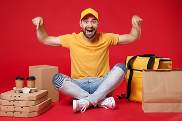 Delivery employee man guy male in yellow cap t-shirt sit at thermal takeaway food bag backpack pizza boxes cups work courier service during quarantine coronavirus covid-19 isolated on red background.
