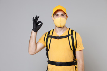 Delivery employee man guy in yellow cap t-shirt uniform face mask gloves thermal food bag backpack work courier service during quarantine coronavirus covid-19 virus isolated on gray background studio.
