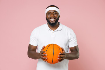 Smiling young bearded african american fitness sports man basketball player 20s in white headband t-shirt playing basketball hold ball isolated on pastel pink color wall background studio portrait.