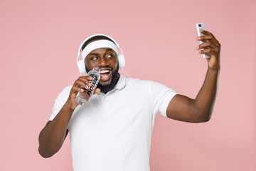 Funny young african american fitness sports man 20s in headband t-shirt listen music with headphones doing selfie shot on mobile phone hold bottle of water isolated on pink background studio portrait.