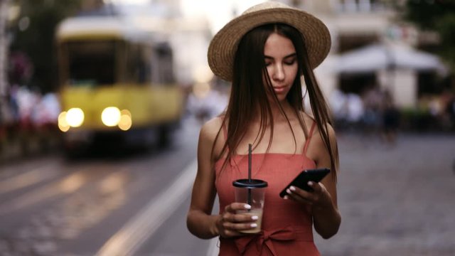 Beautiful young woman in a dress and hat walks around the city and enjoys a warm summer evening. An attractive girl drinks coffee and uses the phone while walking