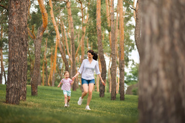 Fototapeta premium Mom and daughter are running in the forest close-up and copy space. The sisters are playing hide and seek in the forest. Mother's day, women's holiday.