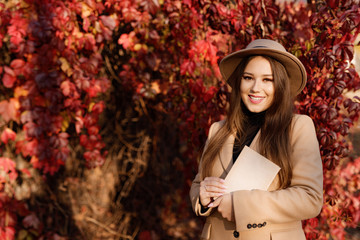 Young woman with a sincere smile having a good time an autumn day. Closeup portrait of charming lady in beige classic coat and hat with book in hands on background of bright autumn leaves.