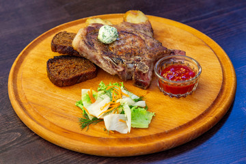 
freshly grilled Tomahawk steak on a round wooden plate with tomato sauce, slices of black bread, lettuce and carrots. Close view