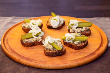 slices of black bread with meat pate decorated with pickles and onions on a round wooden tray. Close view