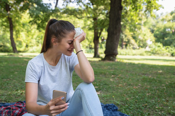 Young sad woman sitting in the park holding mobile phone and crying she got the bad news