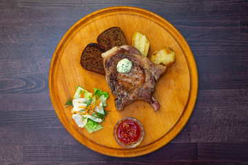 
freshly grilled Tomahawk steak on a round wooden plate with tomato sauce, slices of black bread, lettuce and carrots. Close view