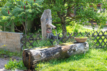 Wooden water bowl and water tap emerging from a chopped tree trunk, carved in the shape of a face